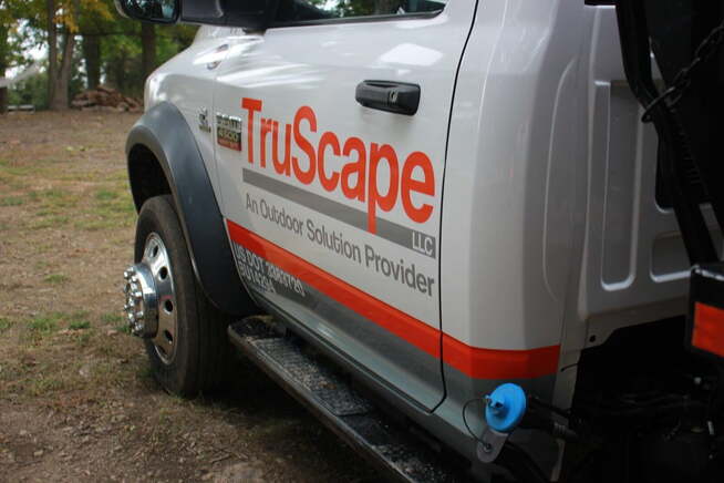 A white commercial truck with "TruScape, An Outdoor Solution Provider, LLC" prominently displayed in red and gray lettering on its side, along with orange and gray accent stripes. The truck is parked on a dirt and grass area, with a large rear wheel and a towing hitch visible. In the background, there are trees and a pile of logs, suggesting a rural or outdoor work environment. A blue object, possibly a tool or equipment, is attached near the hitch.