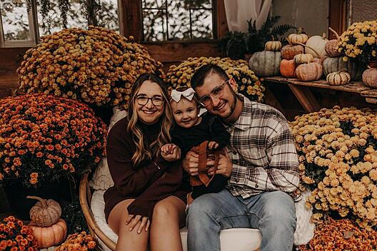 A joyful family of three—two parents and a toddler—sitting together in a cozy, round wicker chair surrounded by vibrant fall decorations. The parents are smiling warmly, with the mother wearing a brown sweater and the father in a plaid shirt and jeans. The toddler, dressed in a dark outfit with a bow in their hair, sits happily between them. The background is filled with colorful autumn flowers, pumpkins, and gourds, creating a festive, harvest-themed setting with large windows and greenery visible behind them.